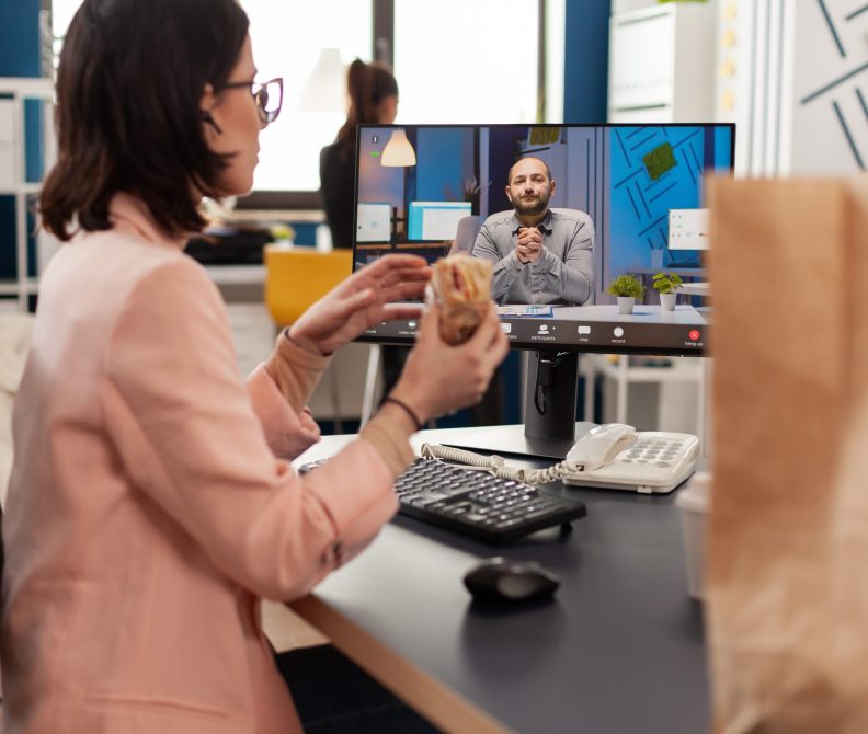 Businesswoman eating delivery takeaway sandwich during online videocall conference meeting discussing with remote coworker. Manager sitting at desk having takeout lunchtime in office