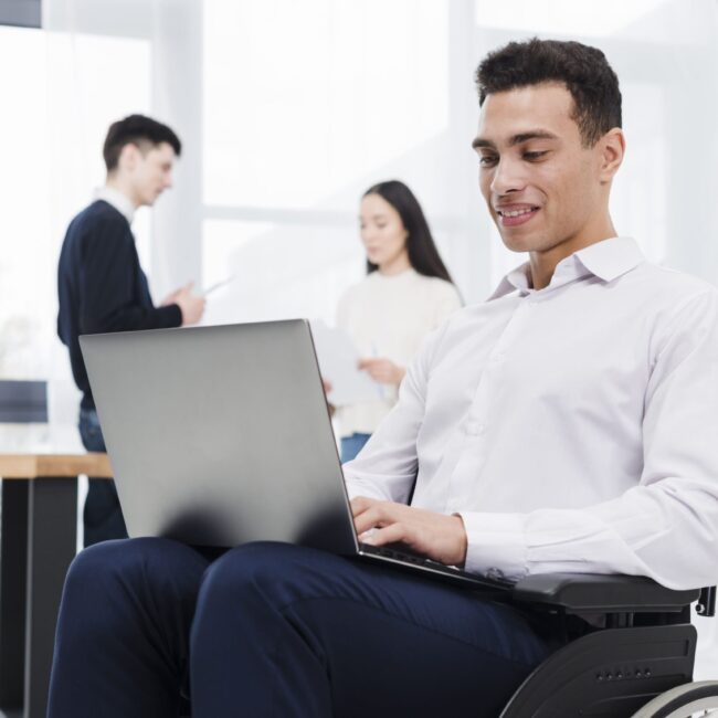 smiling-portrait-young-businessman-sitting-wheelchair-using-laptop-with-his-colleague-background (1)